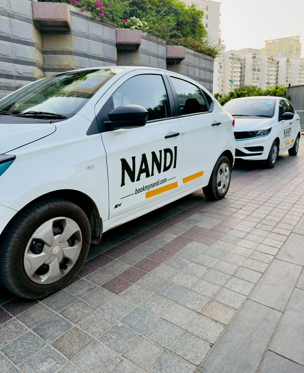Two white Nandi Tata Tigor EV cabs parked nose to tail on a paved driveway, with the Nandi wordmark on the front door.