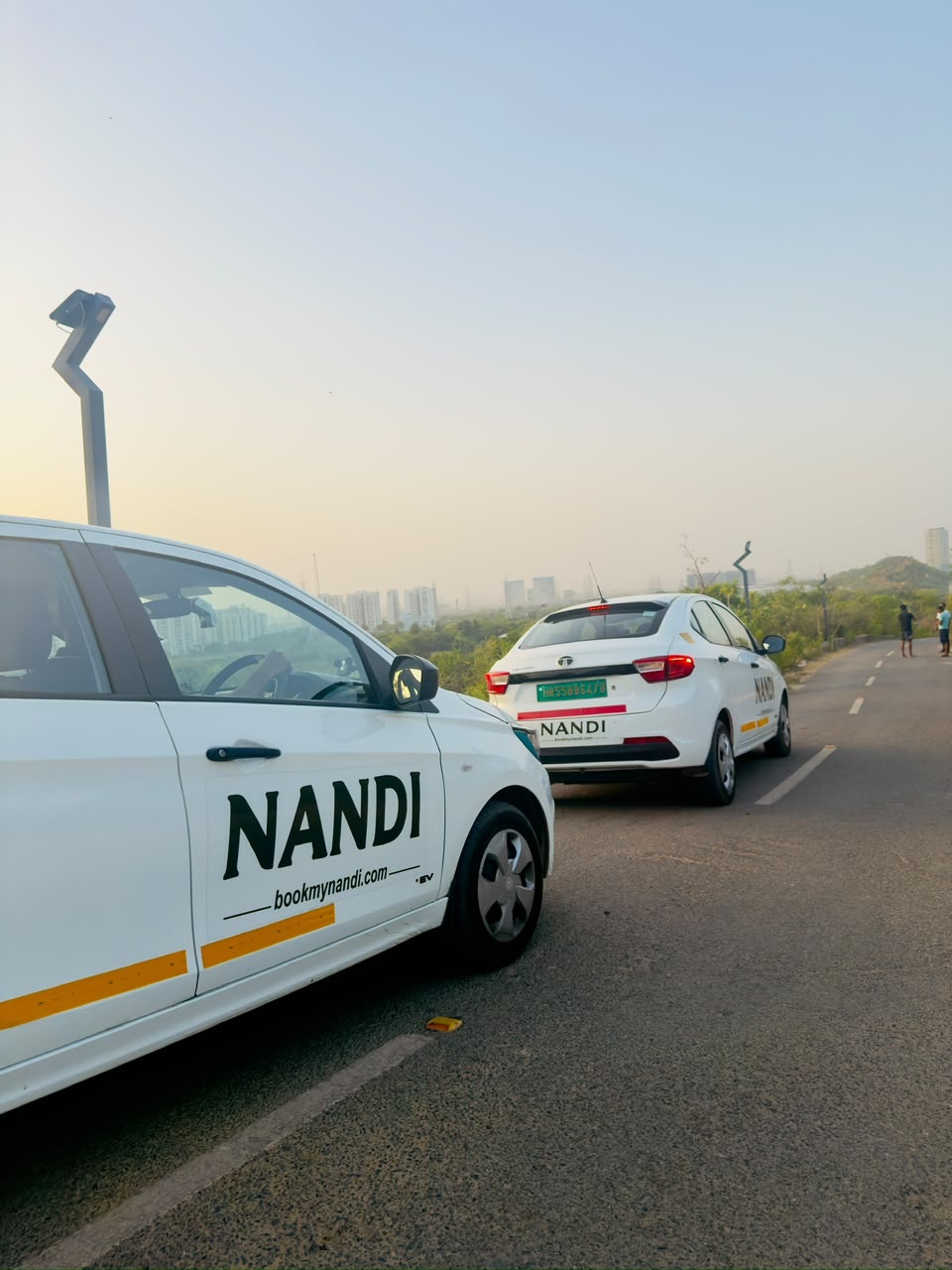 Two white Nandi electric cabs in green and gold livery on a ridge road at sunset, with the Gurugram skyline behind them.